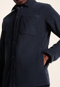 Navy corduroy shirt with a button-up front, chest pocket, and ribbed texture. Features a collar and long sleeves with button cuffs.