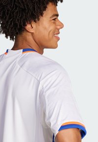 Hvid atletisk trøje med blå og orange detaljer. Den har en rund hals, korte ærmer og en subtil tekstur med et stribet mønster på stoffet.