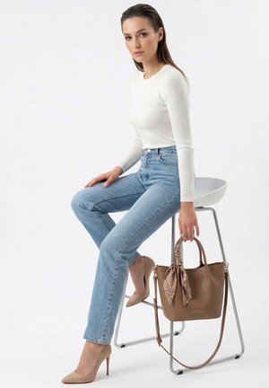 Frau in weißem Pullover und blauen Jeans sitzt auf einem Hocker, hält eine braune Handtasche mit an Henkel gebundenem Schal und trägt beigefarbene High Heels.