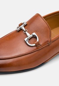 Pantof brown din piele, slip-on, cu un accent metalic în formă de zăbrea de cal și cusături subtile, având o textură netedă și o formă de vârf rotunjit.