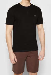 Černé krátké tričko s krátkými rukávy vyrobené z bavlny, s kulatým výstřihem, jemnými švy a malou nášivkou loga na přední straně.