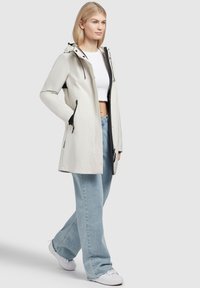 Cappotto impermeabile leggero, beige, con cappuccio, tasche con zip e dettagli neri; abbinato a jeans a gamba larga e sneakers bianche.