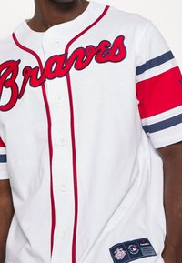 Wit honkbalshirt met rode en marineblauwe accenten, "Braves" script aan de voorkant, knoopsluiting en teammerchandise patch nabij de taille.