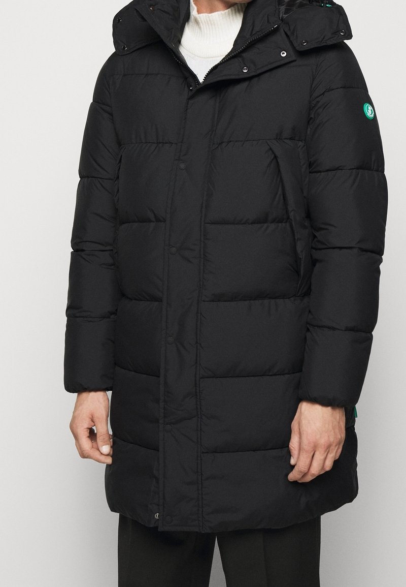 Sort pufferjakke med hætte, har en quiltet design, frontknapper, sidelommer og et grønt logo på ærmet.