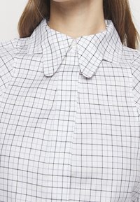 Camisa de color blanca con un patrón de cuadros negros; presenta un cierre de botón en el cuello y suaves pliegues en la parte delantera. Tela de algodón suave.