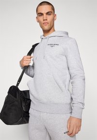 Hombre con sudadera con capucha y pantalones deportivos gris claro de Björn Borg, sosteniendo una bolsa de viaje negra sobre el hombro, de pie frente a un fondo liso.