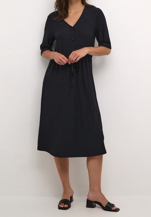 Femme portant une robe noire à mi-genoux avec manches mi-longues, devant boutonné et taille nouée, associée à des sandales noires à talons blocs et bouts ouverts.