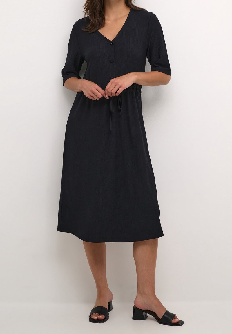 Femme portant une robe noire à mi-genoux avec manches mi-longues, devant boutonné et taille nouée, associée à des sandales noires à talons blocs et bouts ouverts.