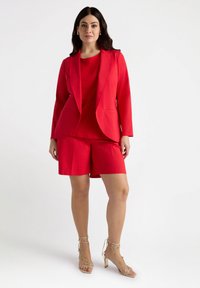 Blazer rojo a medida sobre una blusa y pantalones cortos a juego. La tela es suave con un diseño estructurado. Acentuado con tacones altos nude con tiras.