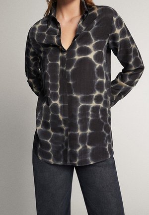 Frau trägt ein langärmliges, schwarz-grau batikmusteres Hemd mit Knöpfen, kombiniert mit dunkelgrauen Hosen, die Hände hinter dem Rücken verschränkt.