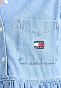 Ljusblå denimskjorta med knappstängning, utrustad med en bröstficka och en "TOMMY JEANS"-logotyp i marinblått, rött och vitt. Volangdetalj i nederkant.