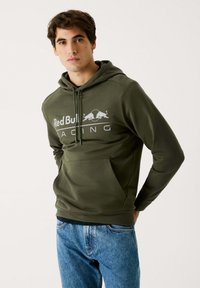Sudadera de color verde oliva con bolsillo canguro, capucha con cordones y un gráfico en blanco de "Red Bull Racing" en la parte delantera. Llevada con jeans azules.