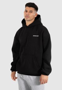 Hoodie noir en mélange de coton, avec une poche avant et une capuche à cordon. Logo blanc brodé sur le côté gauche de la poitrine. Associé à un pantalon de survêtement gris.