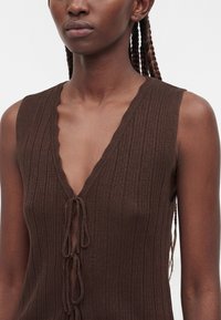 Femme portant un haut en maille marron côtelé sans manches avec un décolleté en V profond et trois nœuds noués sur le devant.