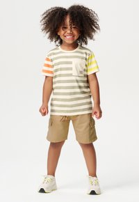 T-shirt a righe per bambini in beige, oliva, arancione e giallo, con taschino sul petto; abbinato a pantaloni cargo color sabbia e sneakers bianche.