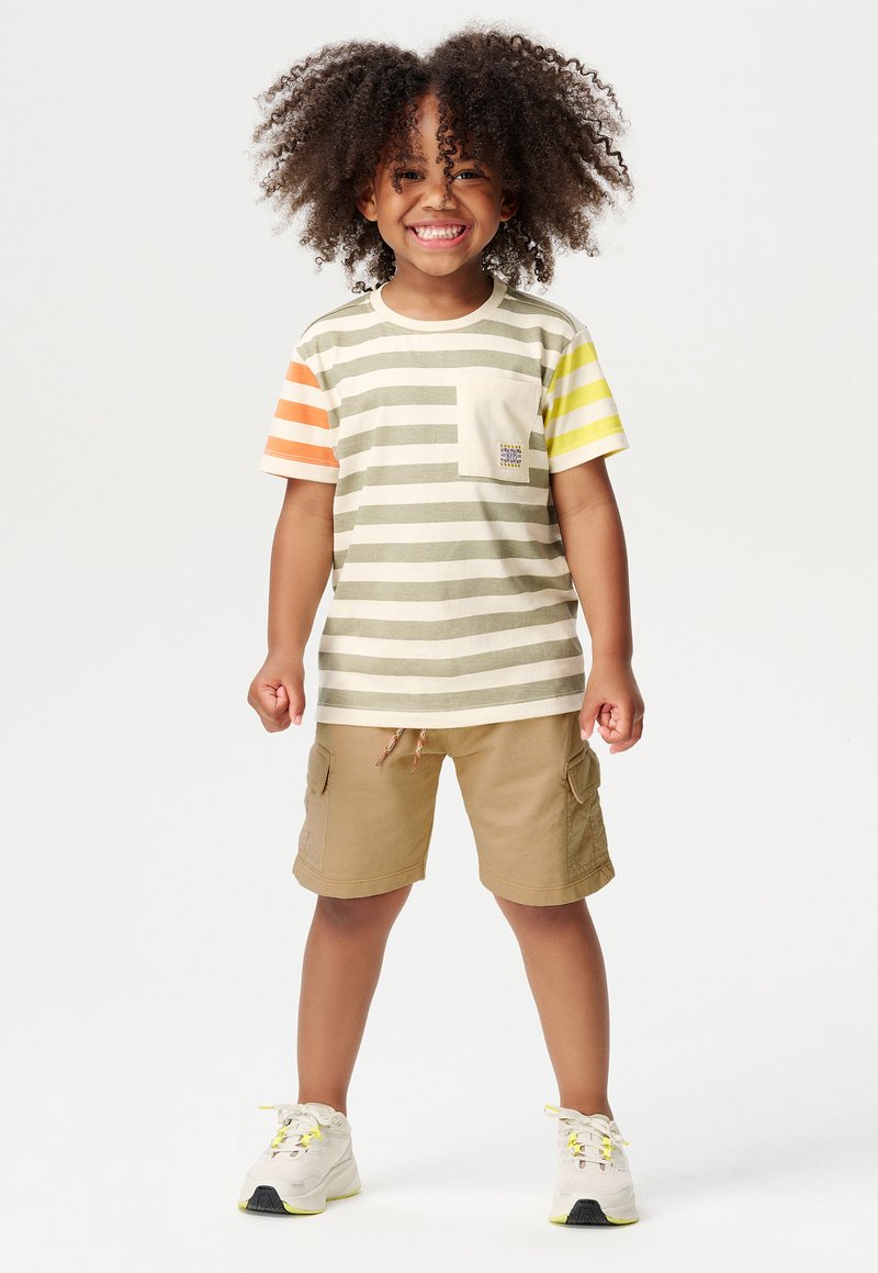 T-shirt a righe per bambini in beige, oliva, arancione e giallo, con taschino sul petto; abbinato a pantaloni cargo color sabbia e sneakers bianche.