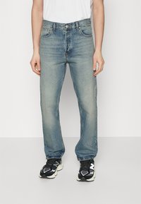 Jeans de denim azul claro con corte recto, cinco bolsillos, un ligero desgaste y cierre de botón y cremallera. Combinados con zapatillas deportivas negras.