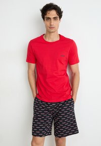 Rotes Baumwoll-T-Shirt mit rundem Ausschnitt, kombiniert mit schwarzen Shorts, die wiederholt weißen und roten "HUGO"-Text aufweisen. Beide Kleidungsstücke haben einen lässigen Schnitt.