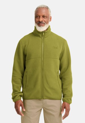 Man met grijze baard die een groene fleecejas met volledige ritssluiting en beige broek draagt tegen een effen achtergrond.