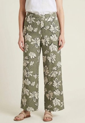 Femme portant un pantalon large vert olive avec un grand motif floral blanc, associé à des sandales slides beige et un haut blanc.