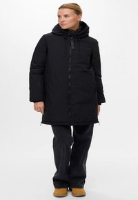 Lang svart puffjakke med glidelås foran, justerbar hette og to sidelommer. Har en glatt tekstur og en lett tilpasset design.