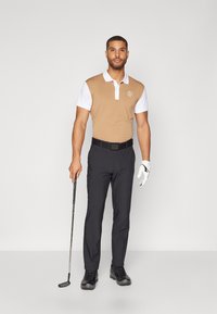 Brūns un balts polo krekls, melnas bikses un golfa apavi. Tur rokā golfa nūju ar kreiso roku cimdiņu, stāvot pret vienkāršu fonu.