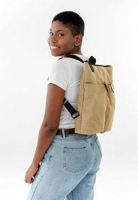 Mochila caqui con detalles en cremallera negra, de forma rectangular, hecha de un tejido duradero, cuenta con dos bolsillos frontales, llevada en la espalda de una persona.