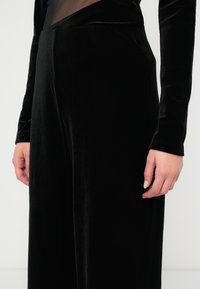 Μαύρο βελούδινο παντελόνι με λεία υφή και άνετη εφαρμογή. Διαθέτει μακριά μανίκια με εφαρμοστές μανσέτες και κομψό, μινιμαλιστικό σχέδιο.