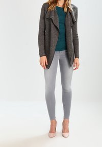 Cardigan pied de poule in bianco e nero con collo drappeggiato, abbinato a una maglia turchese e jeans skinny grigio chiaro. Scarpe con tacchi rosa chiaro completano il look.