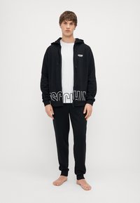 Fekete zipzáros kapucnis felső fehér logóval, párosítva fekete nadrággal. Az összeállítás laza szabású, puha tapintású. A lábak meztelenek.