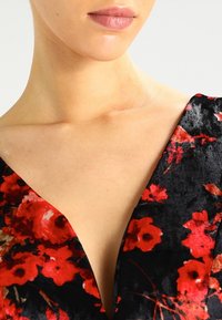 Nahaufnahme des Halses und des oberen Brustbereichs einer Frau, die ein schwarzes Samtoberteil mit rotblumigem Muster und einem tiefen V-Ausschnitt trägt.