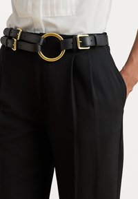 Feketebb bőr dupla öv, nagy kerek arany csatjával, állítható pántokkal és sima textúrával, testreszabott nadrág felett viselve.