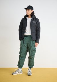 Puffer jacket in black with a hood and logo, white t-shirt, green cargo pants, and gray running shoes with detailed textures and patterns.