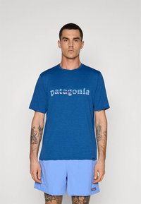 Mėlyna trumpomis rankovėmis pagaminta iš minkštos medžiagos marškinėliai, ant krūtinės su spalvingu „patagonia“ logotipu. Dėvimi su šviesiai mėlynais šortais.