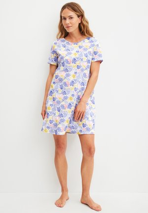 Vrouw staat op blote voeten en draagt een korte mouw, knie-lange jurk met een meerkleurig abstract bladpatroon op een witte achtergrond.