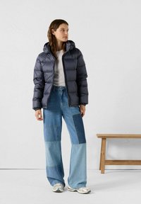 Dikke marineblauwe jas met capuchon, gecombineerd met lichtblauwe wijde patchwork jeans. Model staat tegen een effen achtergrond.