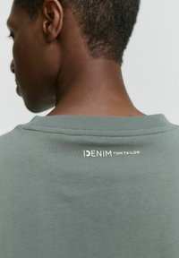 Zelené bavlnené tričko s okrúhlym výstrihom a potlačeným logom značky "IDENIM TOM TAILOR" na chrbte, s hladkou a jemnou textúrou.
