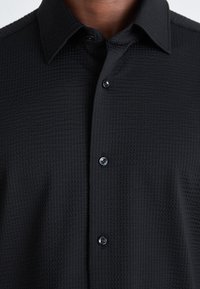 Camisa negra texturizada con cuello puntiagudo y cinco botones negros. La tela tiene un patrón de cuadrícula, que aporta una sutileza de profundidad al diseño.