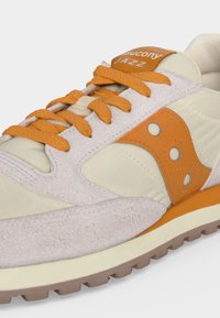 Κοντινό πλάνο μπεζ και πορτοκαλί αθλητικού παπουτσιού Saucony Jazz με πορτοκαλί κορδόνια και λαστιχένια σόλα, που δείχνει λεπτομέρειες υφής από σουέτ και ύφασμα.