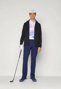 Golf odijelo koje uključuje crnu jaknu, pastelnu grafičku polo majicu, tamnoplave hlače, bijelu kapu i cipele s plavim potplatima. Drži golf palicu.