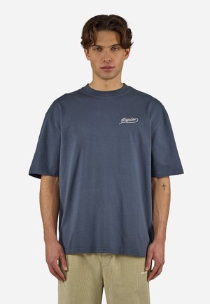 Νεαρός άντρας μεσαίου μήκους καστανά μαλλιά, φοράει μια φαρδιά σκούρα μπλε μπλούζα με λευκή επιγραφή "Pegador" και μπεζ παντελόνι, στέκεται ακίνητος.