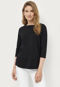 Haut noir à manches longues avec un col bateau. Présente une coupe décontractée et une texture douce. Porté avec un pantalon blanc, les mains placées sur les côtés.