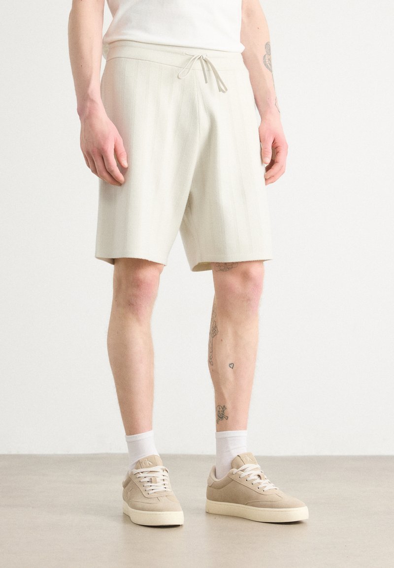 Bermudas texturizadas en tono beige claro con cintura ajustable, con rayas verticales. Combinadas con zapatillas beige y calcetines blancos hasta el tobillo.