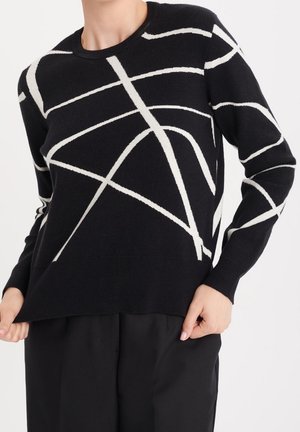 Czarny sweter z długim rękawem z białymi abstrakcyjnymi liniami przecinającymi się, noszony przez osobę trzymającą brzeg, w połączeniu z czarnymi spodniami.