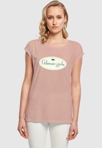 Lys rosa T-skjorte med rullede ermer og rund halslinning. Har en blå oval logo med teksten "Gilmore girls" i midten.