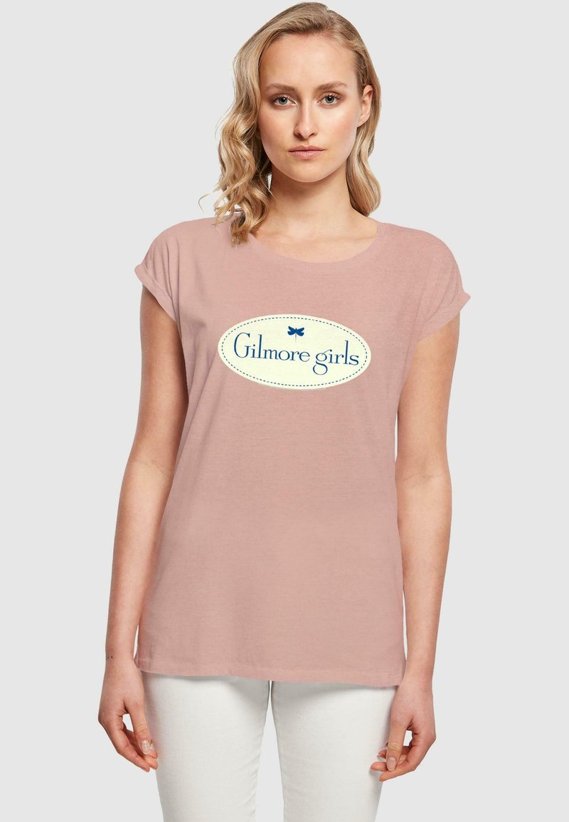 Lys rosa T-skjorte med rullede ermer og rund halslinning. Har en blå oval logo med teksten "Gilmore girls" i midten.