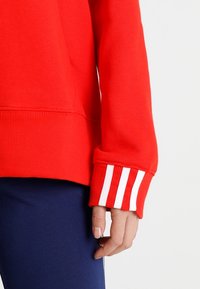 Sudadera roja con dobladillo y puños acanalados, con tres franjas blancas en la manga. Tejido suave y texturizado con un ajuste relajado.