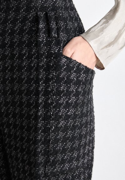 Pantaloni din țesătură tweed neagră și argintie cu un model în carouri, dotati cu un buzunar lateral și o finisare texturată. Mână de personal vizibilă.