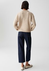 Persona con capelli corti e scuri che indossa un maglione a coste beige, jeans cropped blu scuro e scarpe bianche senza lacci, in piedi davanti a uno sfondo semplice.