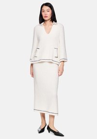 Frau trägt ein weißes, strukturiertes zweiteiliges Outfit mit schwarzem Besatz, bestehend aus einem V-Ausschnitt Langarm-Top und einem Midirock, kombiniert mit schwarzen Absätzen.